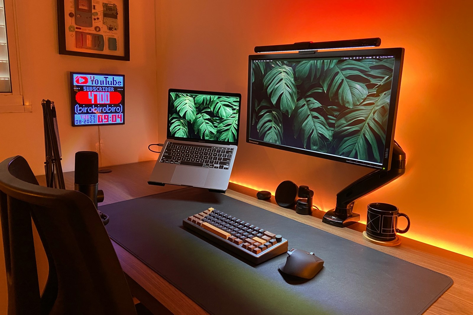 A warm orange-lit home office with a monitor mounted on a black articulated monitor arm, a laptop on a stand, and a large desk mat.