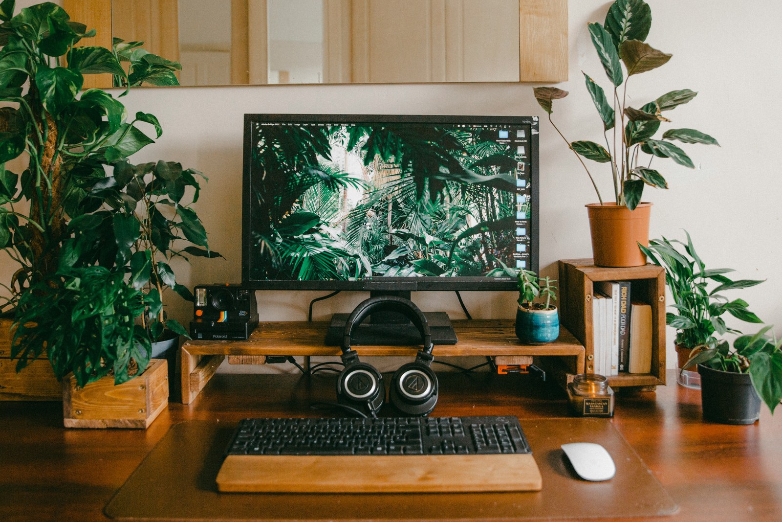 A vibrant home-office desk with a monitor showing a jungle wallpaper, headphones, keyboard, and lush potted plants in warm light.