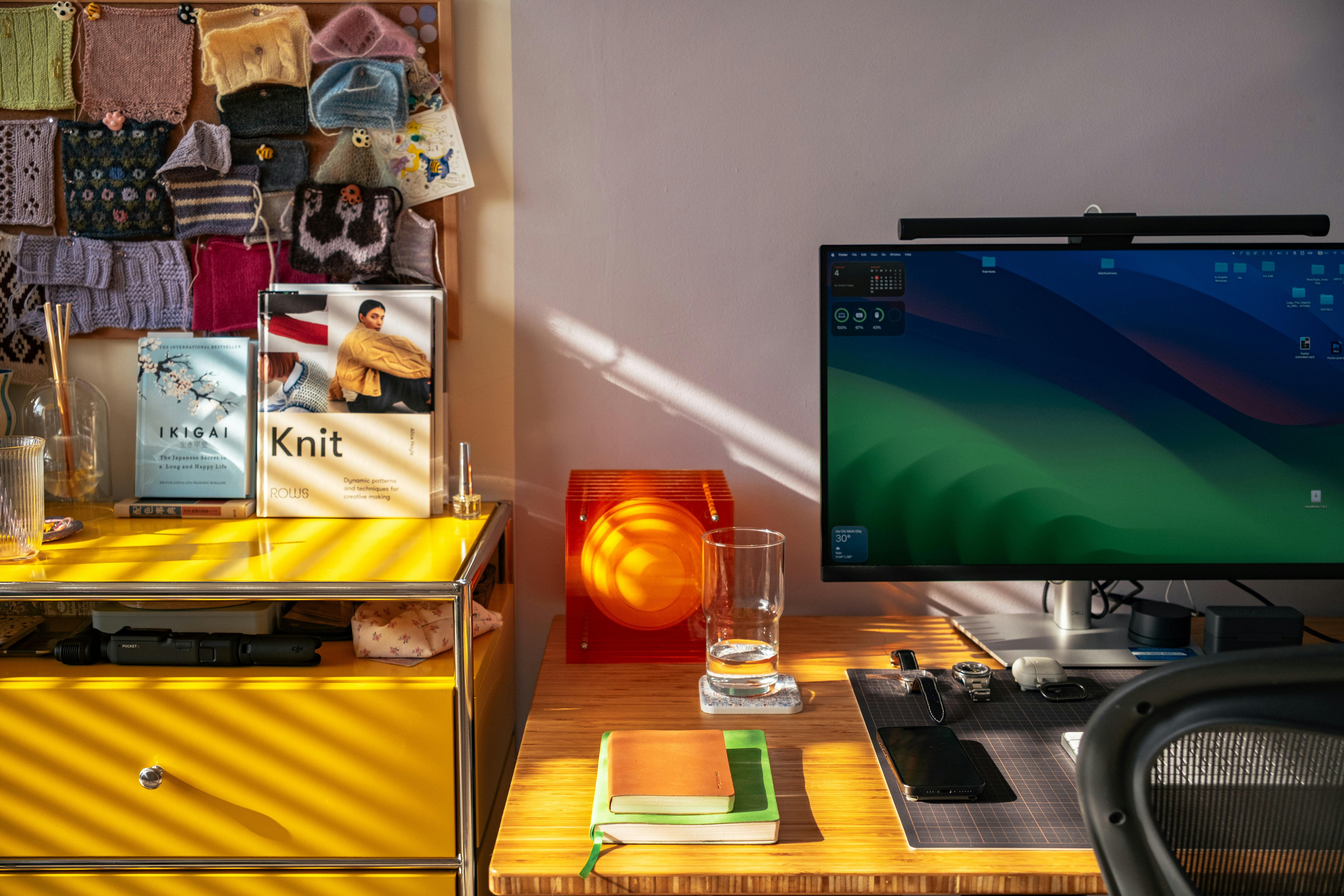 Sunlit home office desk with a computer monitor, notebooks, and a warm orange lamp beside a bright window.