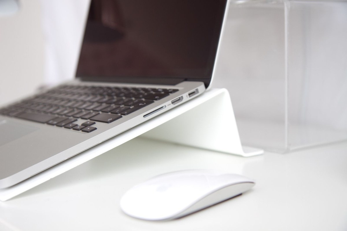 Laptop raised on a white stand beside a wireless mouse on a desk.