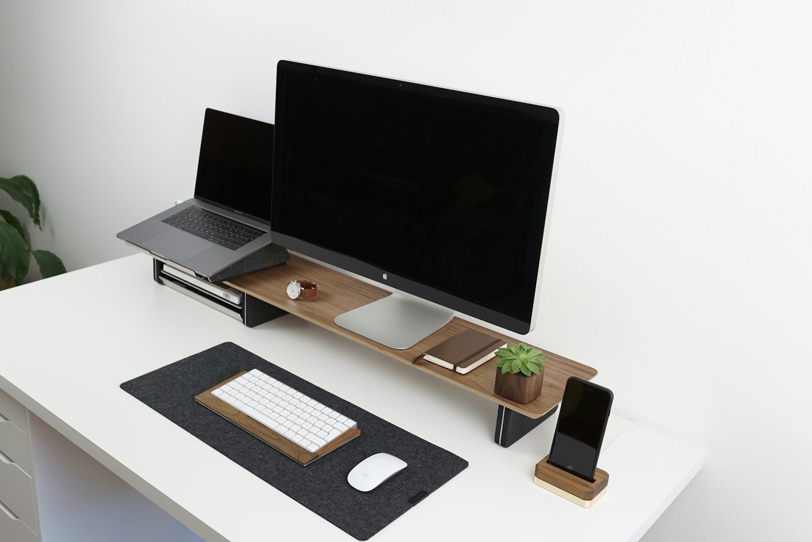 A bright desk setup with a display on a wooden riser, a laptop on a stand, and a felt desk mat with keyboard and mouse.